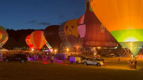 Several colourful hot air balloons lined up with their burners all on at the same time at night. There are colourful lights on the ground, several cars and a large crowd gathered watching to the right.