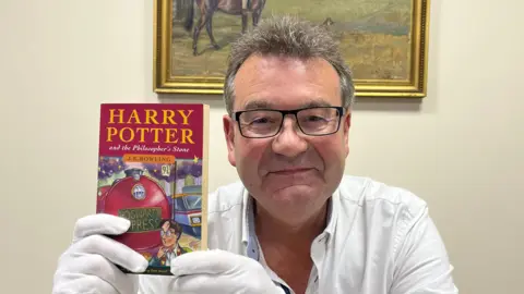 Richard Winterton  Richard Winterton, who has short dark hair and wears black-rimmed glasses, holding the Harry Potter book. He is wearing white gloves and a white shirt. 