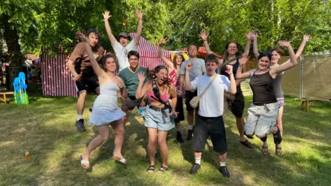 World Jungle A group of young people in summer clothing, jumping up with their hands in the air, on a lawn in dappled sunshine with a red and white striped tent in the background and lots of trees