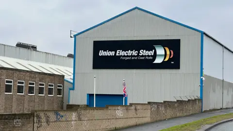 BBC The Union Electric Steel building which is a grey, corrugated building at the side of a side. It has a black sign in the centre which reads 'Union Electric Steel forged and cast rolls.'