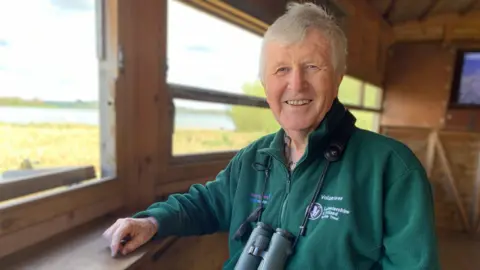 A retired man with grey hair smiles at the camera, with binoculars around his neck. He is sitting in a bird hide and is wearing a green fleece which has 'volunteer' embroidered on it, above a Leicestershire and Rutland Wildlife Trust logo.