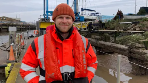 A man in an orange high-vis jacket with a life vest stands with his hands behind his pockets in front of a gangway. He wears an orange hat and is smiling. 