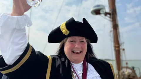 Elliot Deady/BBC The Mayor of Colchester, Lesley Scott-Boutell, raising a glass while wearing her mayoral robes on a barge