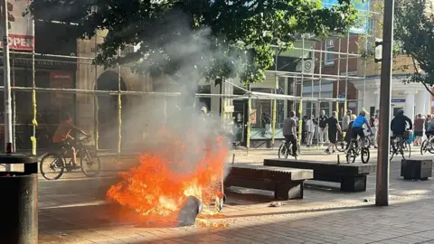 A fire burning from an upturned bin with people riding on their bikes in the background.