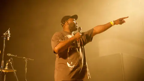 A man on stage in a T-shirt and baseball cap pointing to members of the audience