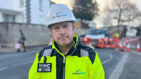 Martin Skyba - a man wearing a yellow florescent jacket with Jersey Water on the front and a white hard hat with the same logo on, a road is behind him blurred.