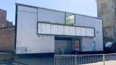 The corrugated facade of a former cinema building which is grey in colour with a green for sale sign above the entrance