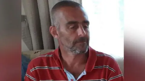 A man with a brown - turning grey - beard with short brown hair sitting on a sofa. He is wearing a red top with white and navy stripes. Behind him is a cream curtain and a window with light shining through. 