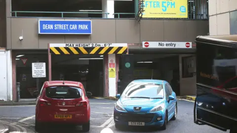 A red and blue car and the edge of a black van parked in front of a multi-storey car park building. It is a large brown building with Dean Street Car Park written inside a blue box. There is a white sign above the entrance which reads: 'Max height 2.0m- 6'7". There is a no entry sign above the exit on the right.