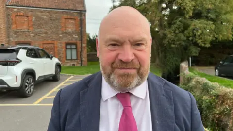 Bill Revans stood outdoors in a car park near a green bushes. He is wearing a dark pink tie and light pink shirt under a navy suit jacket.