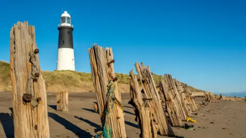 Getty Images Spurn lighthouse, a black and white tower topped with a glass roon, on a bright sunny day beneath blue skies. The photo is taken from the golden-brown beach and in the foreground are a series of weather-worn wooden posts, which stand like sentinels.