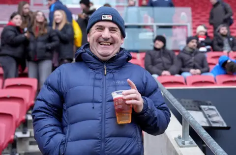 A male fan stands facing the camera at Ashton Gate before a Bristol City Women's match. He has a broad smile and is holding a plastic pint glass. He is wearing a padded blue jacket and a blue bottle hat