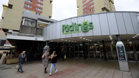 The entrance to the Ridings Shopping Centre, with people walking in and out beneath a curved facade displaying the green “Ridings Shopping Centre” sign.