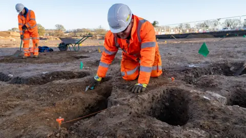 SIZEWELL C Two archaeologists wearing fluorescent orange jackets and trousers, one of them kneeling down, can be seen working on a site of bare earth with holes visible in the ground