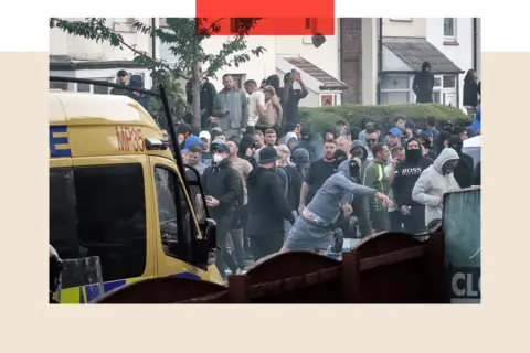 Getty Images A crowd of male protesters behind a police van. One man in the foreground is wearing a black face shield, a blue tracksuit and shorts. He appears to be throwing something.