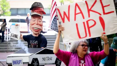Getty Images A model of Elon Musk wearing three hats which say "dork", "Maga", "douche". The model is holding a bag labelled "ketamine" and is in a mock Tesla car where the licence plate says "BNKRUPT". In the foreground is a protestor with a sign which says "wake up".