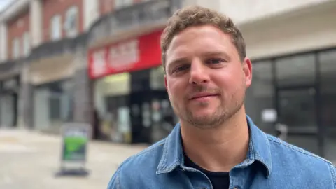 Man with curly hair in a denim shirt in front of a row of shops