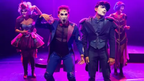 Getty Images four performers on a stage, bathed in purple light. They appear to be in a theatrical performance, possibly a circus or cabaret act, given their costumes and makeup. The costumes are elaborate and theatrical, with one performer in a more traditional, almost mime-like, outfit, while others wear more fanciful, whimsical costumes.