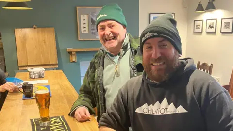 The Fisher's Arms Two drinkers in woolly hats at the bar smiling broadly and ordering pints.