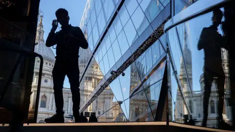 A silhouetted man puts headphones in his ears, standing next to a modern glass building reflecting St Paul's cathedral in the background.