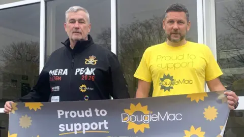 Dougie Mac A man with grey hair in a black sports top and a man with dark hair and a beard in a yellow sports top holding a black banner with yellow flowers and the words proud to support Dougie Mac on it