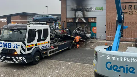 Kaylee Poloczek/BBC A recovery vehicle attempts to extract a burnt out car wedged inside a co-op food store which has also suffered fire damage.