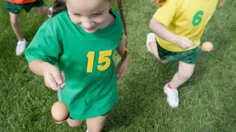 A generic image of sports day at a school. A girl with plaits holds an egg on a spoon and is wearing a green T-shirt with the number 15 on it. Another girl in a yellow t-shirt with a 6 on it is behind also holding an egg and spoon. 