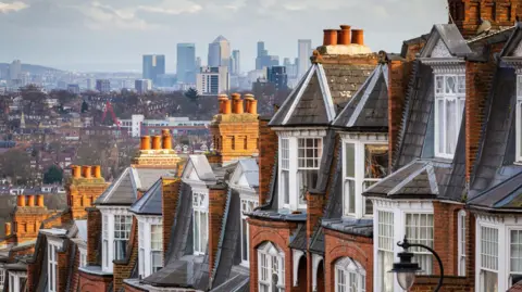 A view of London showing traditional red-brick townhouses with multiple chimneys in the foreground, framed against a distant skyline of modern skyscrapers, including Canary Wharf.