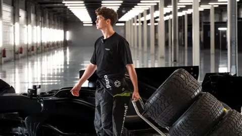 Compound Footwear Alex Witty in a large warehouse-style space in front of a Formula One car as he pulled old tyres behind him