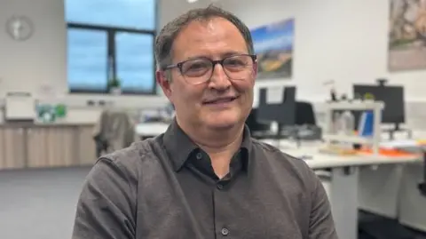 Huw Watkins An office environment with a man seated in the foreground wearing a dark collared shirt. Behind him and slightly blurred, two people are working at desks with computer monitors. The room has white walls, a large window letting in natural light, and office furniture including chairs and storage units. A framed picture is partially visible on the wall to the left. It is a head and shoulders shot of him. 