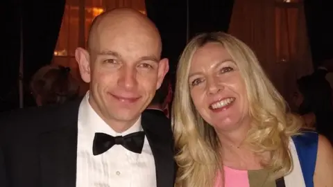 James Williams A bald man wearing a black suit, white shirt and black bow tie sits beside a woman wearing a colourful striped dress with long blonde hair. They are both smiling at the camera.
