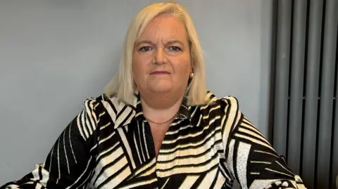 Patricia Lewsley-Mooney A woman is looking directly into the camera. She has blonde, shoulder-length hair and is wearing a black and cream patterned shirt. The wall behind her is grey and to the side there is a horizontal dark grey radiator.