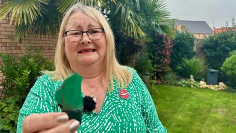 Carol Bailey frowns as she holds a shard of green plastic debris up to the camera. She wearing glasses and a green and white blouse. She is standing in a leafy garden with green grass fringed by neat green shrubs and trees. To her left is a light-brown brick wall. 