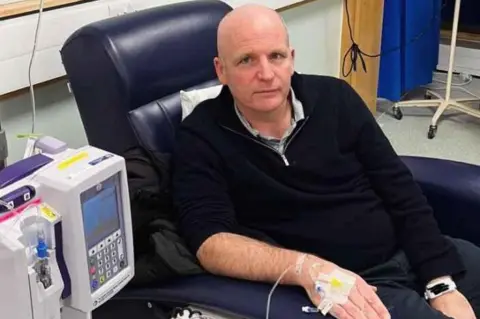 Glenn sitting in a blue chair in hospital where he is undergoing chemotherapy. He is wearing a black zipped top and the medical equipment is visible in the foreground.