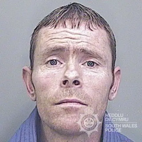 South Wales Police Custody shot of Terry Andrews, aged 36. He has blue eyes and short brown hair. The South Wales Police logo can be seen in the bottom right hand corner. 
