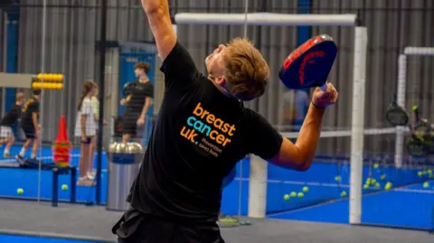 A man playing Padel on a court. He is wearing a black T-shirt and shorts with the words Breast Cancer UK written on the back.