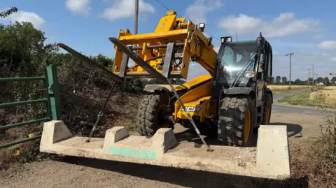 Cambs Police A man is in the cab of a large orange and black JCB digger or tractor, using it to move a large concrete barrier. There are flat fields and telegraph poles along the side of a road in the background