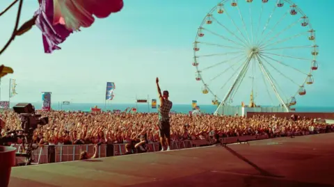 A large crowd of people watching the main stage at Boardmasters festival. There is a large ferris wheel in the background, and the sea can be seen in the distance. A singer is stood on the stage wearing a checkered vest-top and blue shorts, and he is stood with his arm in the air. The crowd are also all stood with an arm in the air and there are flags blowing in the breeze in the background.