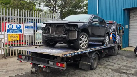 Deputy Andy Cameron A damaged large black SUV style car on the back of a recovery vehicle. The vehicle is parked in what looks like an industrial estate. 
