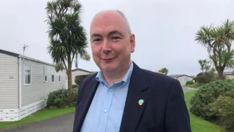 Patrick Langmaid is smiling as he stands outside in the holiday park. There are mobile homes and palm trees behind him. He has short grey hair and is wearing a navy jacket over a blue shirt.