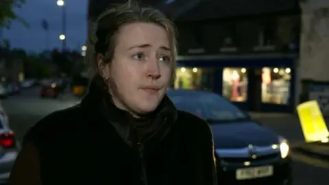 A woman stands next to a road, dressed in a dark coat. A car is just about to drive past her and can be seen over her left shoulder. Three illuminated shop fronts can be seen across the street.