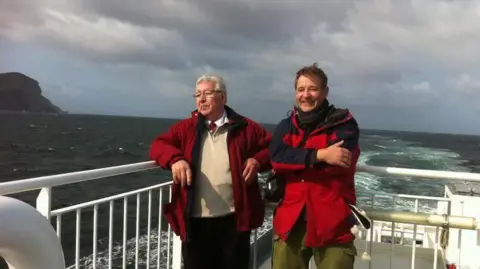 Stuart Flett Two men - Stuart Flett and his dad Rodney - standing on a ferry, with the sea behind them.