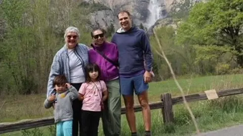 Yorkshire Cancer Research A woman with short grey hair and a blue fleece, stands in front of a mountain with a man, a woman in a purple jacket, and two small children.
