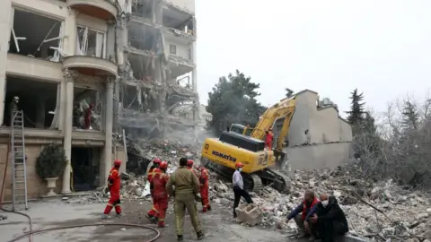 Emergency workers wearing red and grey outfits at the site of a residential building damaged in an attack where a yellow digger is in the rubble; three people wearing civilian clothes, one wearing a red helmet and two face masks, are also in the picture, two of them sitting on the curb edge.