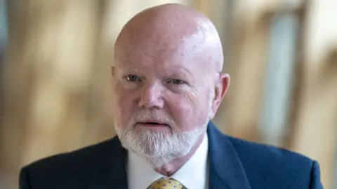 PA Media A bald man with a grey beard walks in the Scottish Parliament