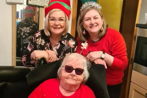 Brenda Young Two women - Brendya young and her sister Janice - wearing Christmas hats, behind their mother Margaret Chisholm who also has Christmas-related items on her head, and dark glasses.
