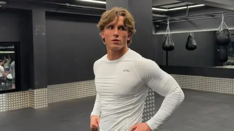 Handout Spencer Clark is standing in a gym space. He is wearing a white long-sleeved T-shirt. 