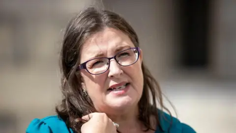 PA Media Armstrong has shoulder length brown hair and is wearing purple framed glasses. She is holding her hair away from her face, and i wearing a blue top.