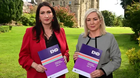 Wakil Pacu Makna Menteri Pertama Emma Little-Pengelly Dalam gugatan merah dan Menteri Pertama Michelle O'Neill dengan setelan abu-abu, meluncurkan kekerasan akhir terhadap kerangka strategis perempuan dan anak perempuan tahun lalu, mereka memegang dokumen di halaman di Stormont Estate dengan Castle di latar belakang