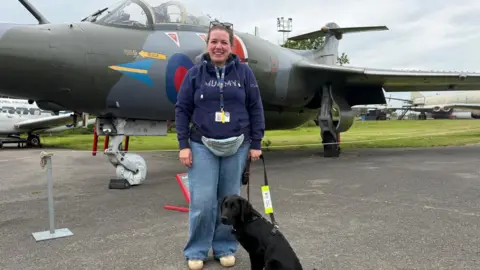 Freya Eady/BBC A woman in jeans and a blue top is standing on tarmac smiling at the camera. Next to her is a black dog on a lead. In the background is a plane that is on display at a museum.